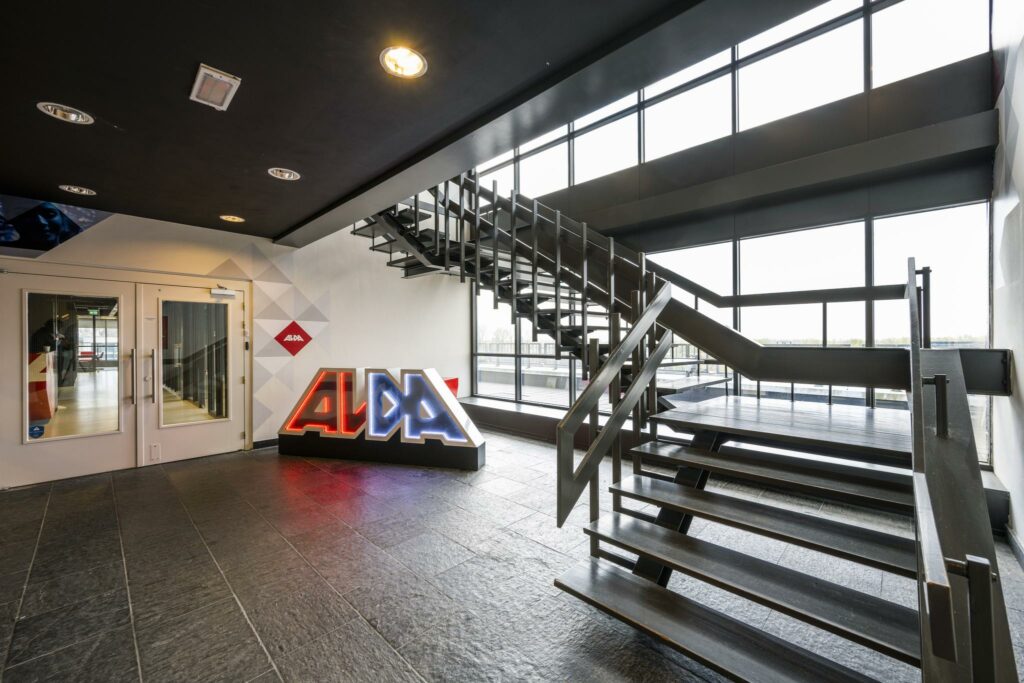 Moderne entreehal met een industriële trap, grote ramen en een verlicht ALDA-logo aan de Anthony Fokkerweg.